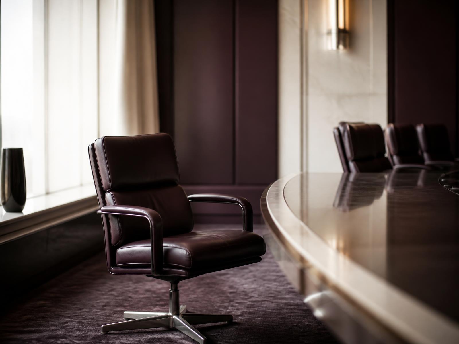An empty leather executive chair pushed back from a polished boardroom table in soft window light